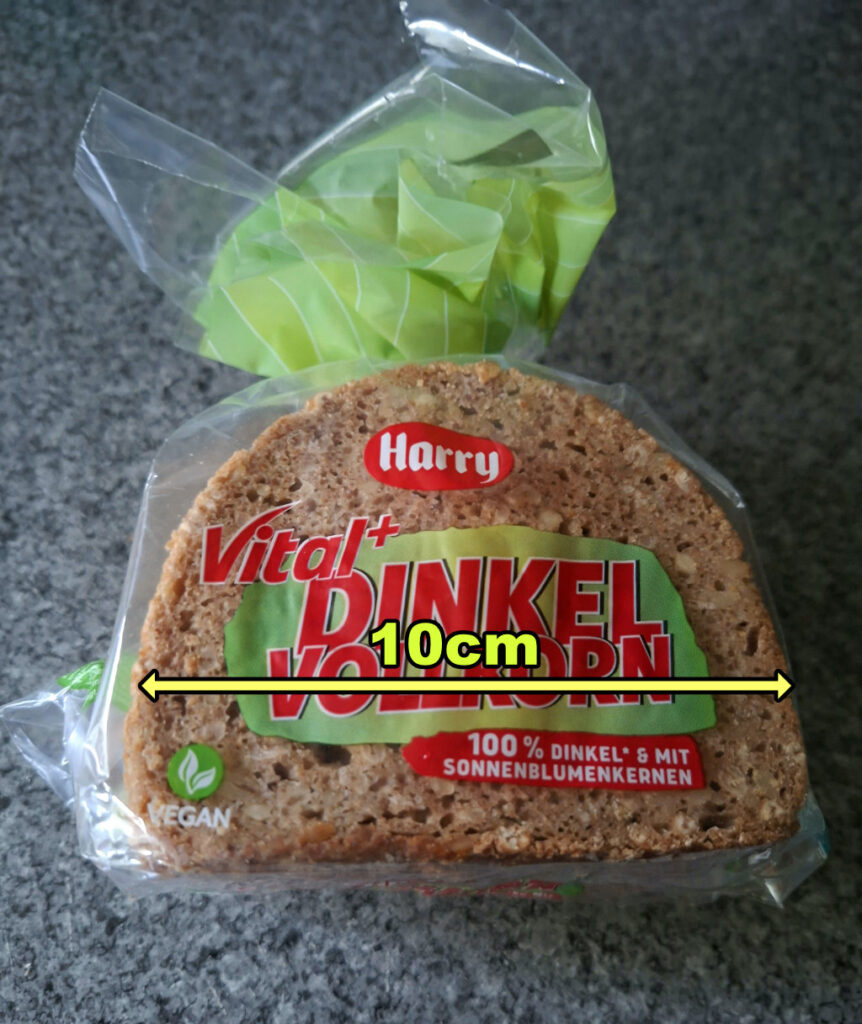 Das Foto zeigt verpackte Brotscheiben der Marke **Harry**, Sorte **„Vital+ Dinkel Vollkorn"**. Die Verpackung ist transparent mit grünen Akzenten. Die Scheibe selbst ist braun, mit deutlich sichtbaren Körnern und den typischen Poren eines Vollkornbrots. Außerdem ist eine gelbe Messlinie im Bild, die eine Breite von **10 cm** markiert.